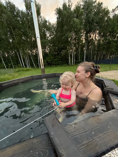 A woman and a young child are in a wooden hot tub filled with water. The woman is wearing a dark bikini and the child is wearing a pink swimsuit. The child is holding a blue water toy. They are outdoors, with trees and grass visible in the background. The sky appears overcast, suggesting it might be late afternoon or early evening. The overall mood seems relaxed and familial. The image is taken in Ryzhkovo, Russia.