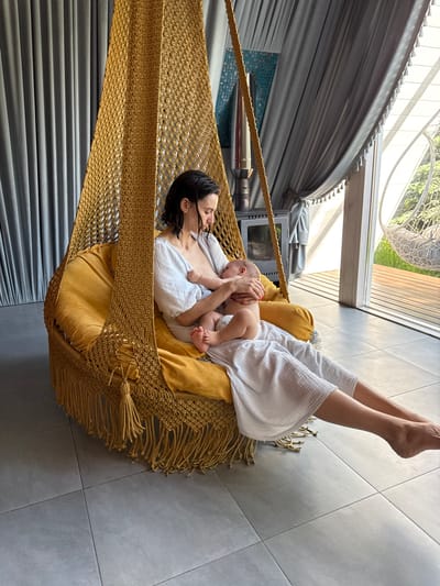 A woman lies in a hammock, holding a baby. The hammock is made of a golden rope material with yellow cushions. The woman is wearing a white robe. The baby is wearing a diaper. The room has gray tiled floors and gray curtains. There is a window behind the hammock, and what appears to be a heater. The location is a spa. There is no definitive proof of the location in the city of Чёрное, Russia.