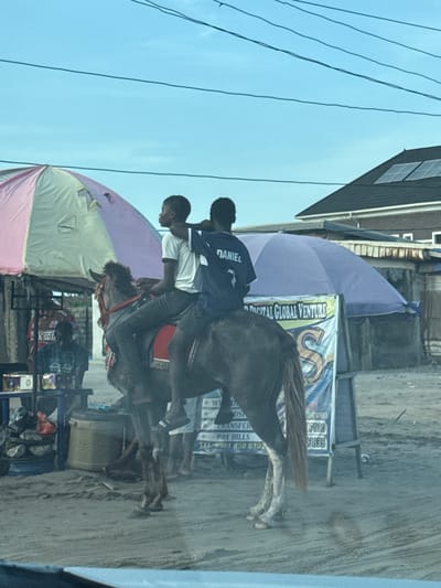 The image shows two young men riding a horse in what appears to be Mopo-Akinlade, Nigeria. The horse is moving on a dirt road. One rider is wearing a white shirt, and the other is wearing a blue shirt with "DANIEL" printed on the back. In the background, there are street vendors under umbrellas and a sign that reads "DIGITAL GLOBAL VENTURE," with phone numbers and payment information. Power lines are visible overhead, and there is a building on the left side of the frame.