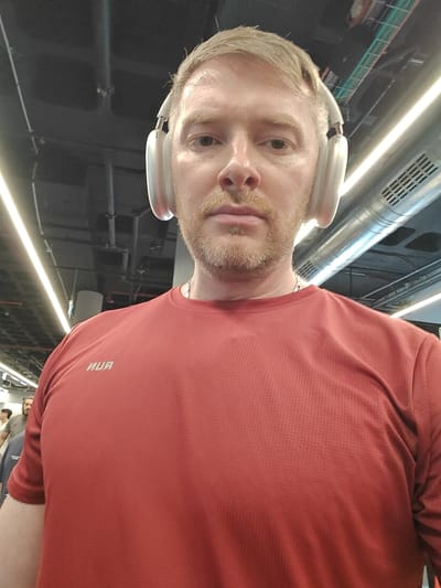 A selfie-style medium close-up photograph taken in the city of Nahariah, Israel, depicts an adult male with light skin, short blonde hair, and a light-colored beard. He wears light-colored over-ear headphones and a reddish-orange athletic shirt with a faint grid pattern, displaying the mirrored word "RUN" on the left chest. A thin silver-colored chain is visible around his neck. His eyes are directed forward, exhibiting a neutral facial expression. The background is an indoor industrial-style environment featuring a dark ceiling with exposed utilities, including pipes, ducts, and linear LED light fixtures. A prominent metallic air duct with horizontal vents is located in the upper right background. Blurred figures of other individuals are discernible in the distant left background.