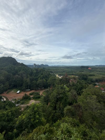 A high-angle, wide shot captures a lush, green landscape under a cloudy sky in Ban Sadet, Thailand. The foreground is dominated by the dense foliage of trees, their leaves a variety of green hues. A dirt road winds through the lower left of the frame, leading to a few small buildings and a white tarp structure. Further into the mid-ground, a cluster of trees gives way to a vista of more trees, with a prominent brown structure, possibly a temple or ruin, situated on a hill. Beyond that, a vast expanse of greenery stretches to the horizon, punctuated by distant, hazy mountains. The sky is filled with scattered clouds, casting a soft, diffused light over the scene.