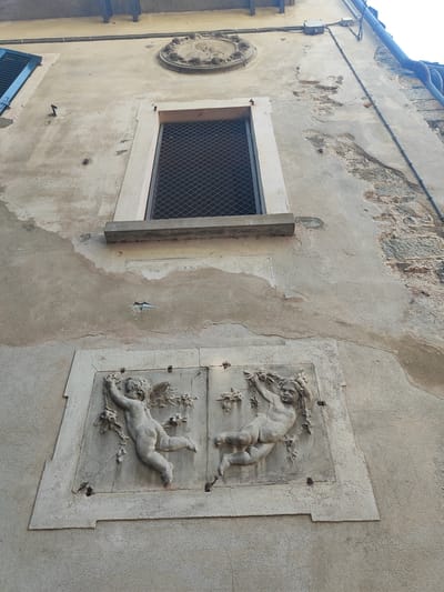 The image shows a close-up, low-angle shot of the exterior wall of a building in Orta San Giulio, Italy. The wall is aged, with patches of peeling plaster and visible stonework. A rectangular window with a metal grate is positioned high on the wall. Below the window, there is a decorative relief carved into the plaster, depicting two cherubic figures holding onto floral arrangements. The cherubs are rendered in a classical style. The scene suggests an old European architectural style, typical of historical towns like Orta San Giulio. The lighting indicates it is daytime, casting natural light on the textured surfaces.