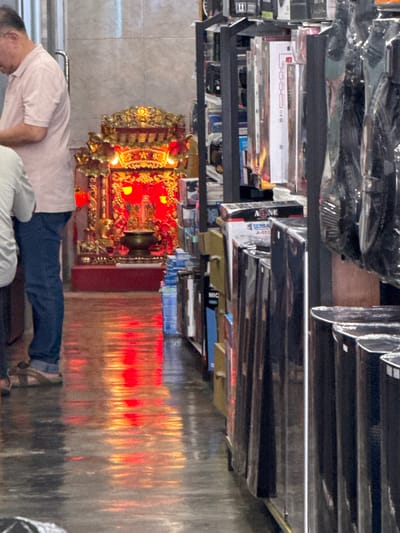 The image depicts an interior scene, possibly a shop, with a man in a light pink polo shirt and blue jeans standing on the left, facing away from the camera. Beside him, to the right, is a vibrant, ornate altar featuring a golden structure with red accents and illuminated by a bright red light. The altar has a dark urn or censer at its base. The floor is reflective, showing a strong red and orange glow from the altar's lighting. In the background and to the right, shelves are filled with various products, some of which are wrapped in plastic. The overall ambiance suggests a place of worship or a shop that incorporates religious elements. The location is identified as Ranong, Thailand.