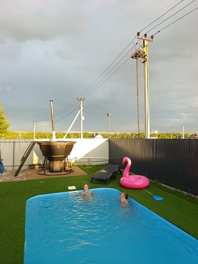 This media file depicts an outdoor scene in a backyard with a swimming pool. Two young individuals, likely children or teenagers, are enjoying themselves in the bright blue water of the pool. One is visible from the chest up, smiling at the camera, while the other is partially submerged. A large, inflatable pink flamingo float rests on the artificial green turf surrounding the pool, with a blue pool skimmer net nearby. A black lounge chair is positioned on the turf, suggesting relaxation.
In the background, a large, dark metallic outdoor cooking station with a chimney is prominent, suggesting a barbecue or similar setup. This is positioned on a concrete pad, with a wooden ladder leaning against it. A white picket fence runs along the edge of the property, beyond which is a dark ribbed fence. The sky is overcast with dark, dramatic storm clouds, hinting at a change in weather, though the lighting suggests it is late afternoon or early evening, with some sunlight breaking through. Power lines and utility poles are visible against the stormy sky, indicating a semi-rural or suburban setting. The overall atmosphere is one of leisure and fun, despite the impending weather. No visible text is present in the image.