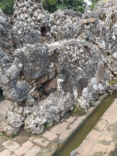 The image shows a large, grey, rocky structure, possibly a man-made cave or decorative rock formation, at the Keraton Kasepuhan palace in Cirebon, Indonesia, located at the given coordinates. The rock is formed from smaller stones and pebbles embedded in a matrix of cement or mortar. A long, narrow channel filled with murky water runs along the bottom of the image and alongside the base of the rocky structure. The channel appears to be made of several flat stones laid in a line. Vegetation grows sparsely around the edges of the water channel and at the base of the rock formation. There are no people present in the scene.