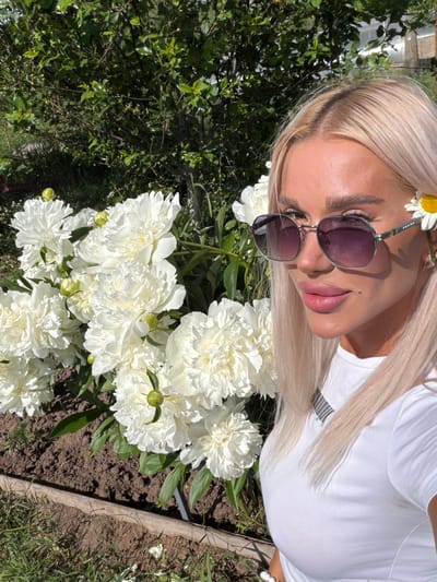 A blonde woman wearing sunglasses and a white t-shirt stands in front of a bush of white peonies. A daisy is tucked behind her right ear. The woman is looking at the camera, and her lips are slightly parted. The background is a blur of green foliage and a wooden border. The scene appears to be outdoors in a garden. The location is identified as Aryshkhazda, Russia.