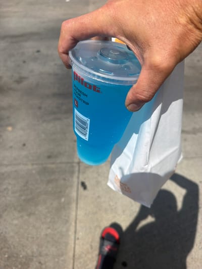 A person's hand is holding a large, clear plastic cup filled with a bright blue beverage. The cup has a clear plastic lid with a circular opening. The logo "Pilot" is visible in red on the side of the cup, along with text that reads "REFRESH YOUR ROADTRIP." A white paper bag with orange lettering is tucked under the cup, held by the same hand. The background is a paved surface, likely a sidewalk or road, with shadows cast by the sun. A shoe with a black band and red accents is visible in the lower part of the frame. The scene appears to be outdoors during the day. The location is the city of Hayti, United States of America.