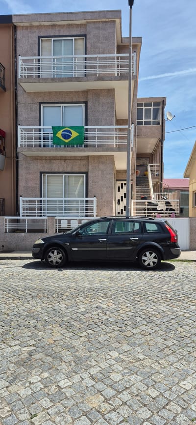 A dark station wagon is parked on a cobblestone street in front of a multi-story building. The building has a facade of mottled brown tiles and features several balconies with white railings. A flag of Brazil is hanging from the railing of the second-floor balcony. A set of outdoor stairs is visible on the right side of the building, leading up to an entrance. The sky is blue with some wispy clouds. The scene appears to be in a residential area of Vila do Conde, Portugal.