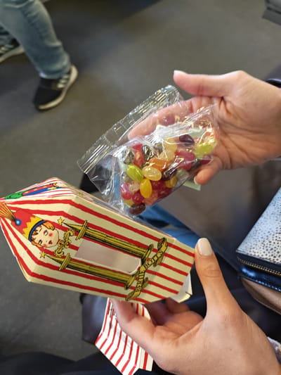 A person holds a clear bag of Bertie Bott's Every Flavor Beans above an opened Bertie Bott's box, with another person's legs visible in the background. The location is Mid Sussex, United Kingdom.
