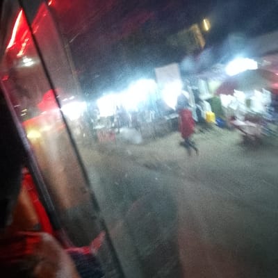 A blurry, nighttime scene appears through a vehicle window. Bright lights illuminate a market area with stalls and people. A figure in a red skirt walks or runs. The view is obscured by the window's reflection and low light, creating a hazy and unclear image. Red lights are visible within the vehicle.