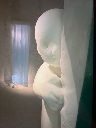 This image depicts an interior scene within an ice structure, characteristic of the ICEHOTEL in Kiruna, Sweden. A large, stylized sculpture of an infant or young child, carved from compacted snow or opaque ice, occupies the foreground on the right. The figure is light-colored, featuring smooth, rounded contours, and appears to be leaning against or emerging from a larger, vertical ice column, with its right arm and hand visible. The child's face is rendered with minimalist features, looking downwards and slightly to the left. In the background, to the left, a tall, translucent ice sculpture resembling draped fabric or a curtain is present, illuminated internally or from behind with a distinct pale blue light. The floor and surrounding environment are composed of white snow or ice, with subtle textural variations. The scene is illuminated by artificial light, creating soft highlights and shadows that emphasize the sculpted forms. No human individuals are present in the frame.