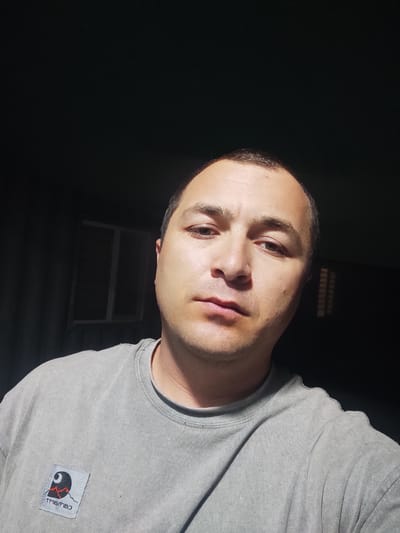In a dimly lit interior space, a man is pictured from the chest up, looking directly at the camera. The man has short, dark hair, brown eyes, and a neutral expression. He is wearing a light grey t-shirt with a small square patch on the left side of his chest. The patch features a black logo with red mountains and some text below it. The background is dark and appears to be a wall with some indistinct features. There is a window or opening visible on the left side of the background, with a light shining through it, casting shadows. The lighting suggests the image was taken at night or in a dark room, with a single light source illuminating the man's face and upper body. The setting is consistent with a residential area in Shaydon Town, Tajikistan.