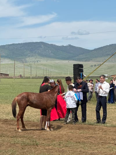 A group of people are gathered around a brown horse in a field with rolling green hills in the background under a partially cloudy blue sky. In the foreground, a man in a white shirt is clapping, facing a man in a black suit who is holding a medal and ribbon. A young boy in a white shirt with blue trim is standing between them, looking at the medal. To the left, a man in a black shirt and a woman in a red dress are standing near the horse. Further back, a stack of black speakers is visible, as well as tents and fencing. The overall scene appears to be an outdoor award ceremony or event, possibly in Orongoi, Russia, given the mountainous terrain and style of dress.