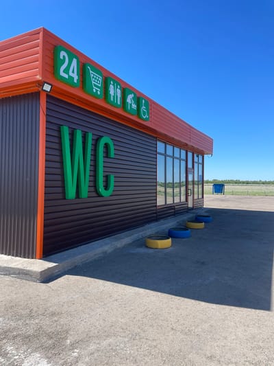 The image shows a building in Arshaly, Kazakhstan. The building has an orange and brown exterior with the sign "WC" and other symbols indicating services such as shopping and 24-hour availability. Blue and yellow painted tires are lined up next to the building. The sky is clear and blue, and there is a field visible in the background.