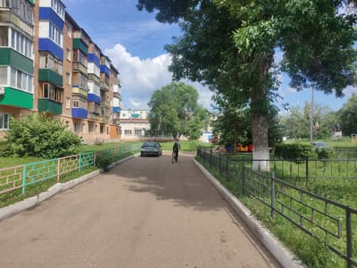 A low-angle, wide shot captures a street scene in Salavat, Russia, on a sunny day with scattered clouds. A multi-story apartment building, characterized by its brick facade and colorful balconies (green, blue, and white), dominates the left side of the frame. A paved pathway, bordered by a low fence with green and pink horizontal bars, runs along the building. Further down the pathway, a dark-colored sedan is parked, and a person on a bicycle is riding away from the viewer. To the right of the pathway, a mature tree with dense green foliage casts shadows, and a black metal fence encloses a grassy area. In the background, other buildings and trees are visible, under a bright blue sky.