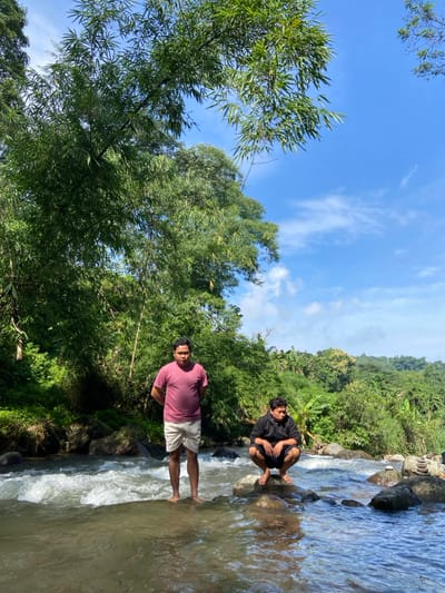 Two men stand on rocks beside a stream. One man in a dark shirt sits on a rock. The other in a pink shirt stands in the water with his legs visible. Lush green vegetation lines the stream bank. Trees extend into a blue sky with some clouds. Water rushes over the rocks.