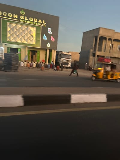 The image depicts an outdoor street scene in Kofar Ruwa, Nigeria, during what appears to be dusk or early evening.
On the left, a prominent commercial building features a large sign that reads "IBCON GLOBAL RESOURCES LTD." Below this, additional text, partially obscured, appears to include "Engineering and General Merch...". The building's facade is adorned with several colorful, irregularly shaped lights or decorative elements, and a large black water tank is visible near its base.
In front of this building, a group of approximately eight to ten individuals are engaged in communal prayer, kneeling and prostrating on the ground with their backs to the viewer. They wear various casual garments, some with head coverings. A white truck is parked behind the praying group.
Further down the street, another multi-story concrete building stands, with two yellow auto-rickshaws visible nearby. A lone individual is seen standing upright closer to the background, near the roadside.
To the right, a paved road is visible, demarcated by a curb painted with distinct black and white stripes. The scene captures a moment of religious observance within a bustling urban environment.