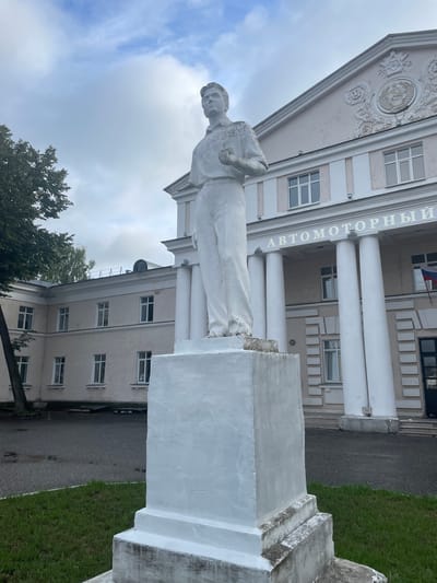 The image shows a white statue of a young man standing on a rectangular pedestal in front of a light-colored building. The building has several windows and large white columns, and there is Russian text above the columns that reads "АВТОМОТОРНЫЙ" (Automotorny). A Russian flag is visible on the right side of the building. The statue is in the foreground, and the building occupies the background. The area around the statue appears to be an outdoor plaza or courtyard, with some grass and a paved surface. The sky is overcast. This scene is located in Zavolzhye, Russia.