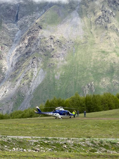 In a grassy mountain meadow in Gergeti, Georgia, a blue and white helicopter with the registration "4L-GSG" sits on the ground. Three individuals are present near the helicopter. One person, wearing a high-visibility yellow jacket, stands by the open door of the helicopter. Another person, wearing a dark shirt and blue jeans, stands facing away from the helicopter. A third person, in a blue shirt and dark pants, stands with their back to the helicopter, looking towards the mountains. The background is dominated by a steep, rocky mountain slope covered in patches of green vegetation. A line of trees borders the meadow in the middle ground. The overall scene depicts a helicopter landing or preparing for departure in a remote, mountainous environment.