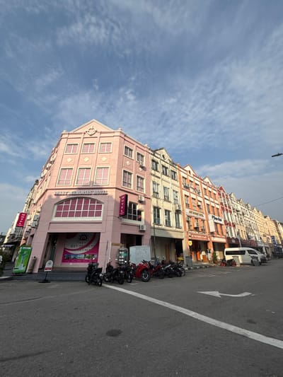The image captures a daytime street scene in Malacca City, Malaysia, featuring a row of multi-story shophouses under a clear blue sky with scattered cirrus clouds. The prominent building on the left, located at what appears to be a street corner, is the "Sweet Memories Hotel," a four-story structure with a pink facade, a gabled roof, and numerous windows, some framed in pink. A red vertical "HOTEL" sign is mounted on its right side, and a large banner with contact information ("017-2900 535") is displayed on the ground floor. A green vending machine is visible to the immediate left of the hotel.

Extending to the right, a continuous line of three- to four-story shophouses follows the street's curve. These buildings display varied architectural designs and colors, including cream, orange, and white facades, with visible signs for businesses such as "AL KHAIR BAKERY & CAFE" and "Tiga Hotel." Multiple external air conditioning units are attached to the upper levels of these buildings.

The foreground consists of an asphalt road featuring a solid white lane marking and a white arrow indicating a right turn. Approximately ten motorcycles are parked in a cluster on the street or sidewalk area in front of the initial buildings. Further down the street, a white passenger van is parked kerbside. No individuals are visible within the frame. The lighting indicates direct sunlight, with shadows falling towards the left, consistent with a sunny day.
