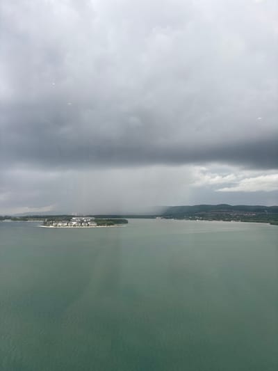The image depicts an elevated view of Falmouth Bay in Falmouth, Jamaica, under heavy, overcast skies. The upper two-thirds of the frame are dominated by a dense, dark gray cloud layer, from which a distinct curtain of precipitation is visibly descending in the mid-left area. The lower half of the image features a calm, teal-colored body of water. In the mid-left, a light-colored peninsula or small island extends into the bay, hosting a cluster of approximately six to eight multi-story buildings with light-colored facades and roofs, surrounded by green vegetation. Further in the distance, along the left shore, taller, possibly industrial or port structures are discernible. A range of verdant hills and mountains forms the distant horizon across the bay, with scattered structures visible along the distant shorelines. No human figures or active vehicles are identifiable.
