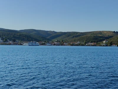 The image shows a body of calm blue water in the foreground, with a coastal town and hills in the background. A large white ferry boat is docked in the harbor, with other smaller boats visible. The town consists of several buildings with red-tiled roofs, nestled against the green, tree-covered hills. The sky is clear and blue. The Municipal Unit of Pelasgia, Greece is a coastal area, and this image likely depicts a view from the sea towards one of its harbors or coastal villages.