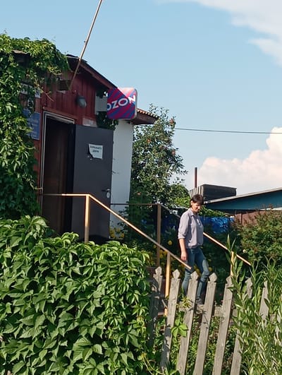 The image depicts a residential setting in the village of Novokopylovo, Russia, on a clear, sunny day. In the foreground, a weathered wooden fence borders a lush green garden. A woman, dressed in a light blue collared shirt and blue jeans, is descending a set of wooden stairs. The stairs lead up to a small, red wooden building with a white facade. A sign with the "OZON" logo, featuring red and blue stripes, is mounted on the building. A dark gray door is visible on the building's exterior, with a smaller sign reading "ЗАПАСНОЙ ВЫХОД" (Emergency Exit) affixed to it. The area is characterized by dense vegetation, including vines climbing the building and surrounding trees, suggesting a rural or semi-rural environment. In the background, other structures and trees are visible under a bright blue sky with scattered white clouds.