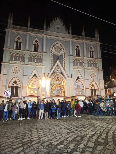 A large group of people stands in front of a pale blue gothic church in El Cisne, Ecuador, at night. The church is illuminated and features intricate architectural details, including spires, arched windows, and decorative trim. The crowd appears diverse in age and attire, and they are facing the church, possibly as part of an event or gathering. The foreground consists of a cobblestone street. A "no parking" sign is visible on the left. Buildings and other structures can be seen to the right. Overhead wires crisscross the sky.
