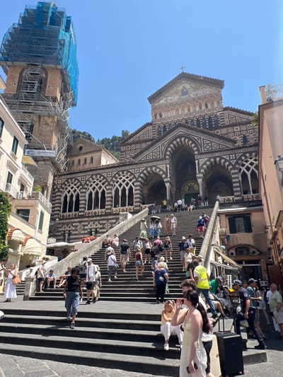 This image captures a wide-angle view of a bustling public square with a grand cathedral as the backdrop, situated in Pogerola, Italy. The cathedral, a prominent structure, is adorned with intricate geometric patterns and features a large arched entrance. A tall bell tower, partially obscured by scaffolding and blue netting, stands to the left of the cathedral. Numerous individuals are seen ascending a wide set of stone stairs leading to the cathedral. They are dressed in casual summer attire, indicative of a warm, sunny day. Some people are walking, while others are seated on the steps, resting or observing. Among the crowd, there are individuals carrying luggage, suggesting they might be tourists. A person in a bright yellow vest, possibly a worker or guide, is visible on the stairs. The environment is lively, with a sense of movement and activity as people navigate the steps and the plaza. The architecture and atmosphere suggest a popular tourist destination.