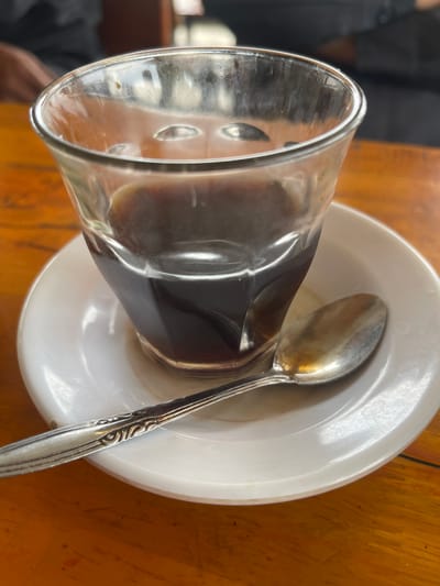 In Julikeude Dua, Indonesia, a close-up shot captures a glass of dark liquid, likely coffee, resting on a white saucer. A silver spoon with an ornate handle lies on the saucer, partially beneath the glass. The scene is set on a wooden table with an orange hue. The background is blurred, showing hints of people.