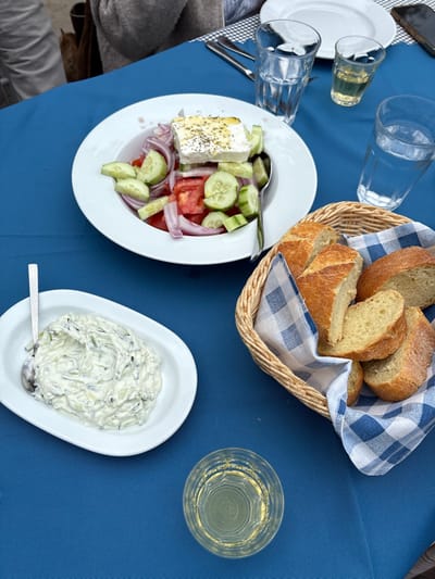 This image depicts a dining table set for a meal at a location within the Municipal Unit of Loutraki - Perachora, Greece. The table is covered with a blue tablecloth.
In the upper center, a white bowl holds a Greek salad (Horiatiki Salata) consisting of large chunks of red tomatoes, sliced cucumbers, and red onions, topped with a substantial block of feta cheese sprinkled with dried herbs, likely oregano, and olive oil. A silver serving spoon rests in the salad.
To the lower left, a white oval dish contains tzatziki, a creamy white dip with visible cucumber shreds, with a silver spoon resting within it.
On the right side of the table, a light brown wicker basket lined with a blue and white checkered (gingham) cloth napkin holds several slices and chunks of light-crusted bread.
Beverages include two tall clear glasses of water: one positioned above the Greek salad, and another to the right, behind the bread basket. Additionally, two smaller clear glasses contain a light yellow liquid, possibly wine or ouzo; one is situated between the two water glasses, and the other is in the lower center of the frame.
In the upper right background, two empty white plates are stacked. Adjacent to them, a dark-colored mobile phone is partially visible. A silver fork and knife are placed above the Greek salad bowl on the upper left. Parts of two individuals are visible in the background: an arm wearing a grey sleeve on the upper left, and another person's arm or clothing in the far upper left, indicating at least two people are seated at the table.