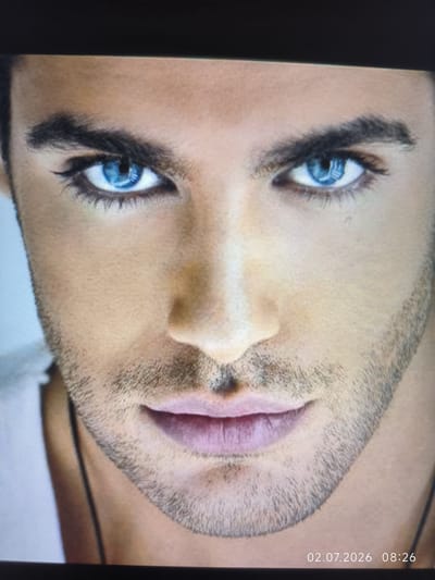 The image is a close-up, frontal portrait of a man's face. He has striking blue eyes with dark eyeliner and full, pink lips. His eyebrows are dark and well-defined, and he has a light stubble beard. The background is out of focus and appears to be a light, neutral color. A timestamp in the bottom right corner reads "02.07.2026 08:26". There is no indication of the location being in Kattasu, Uzbekistan or any other specific city.