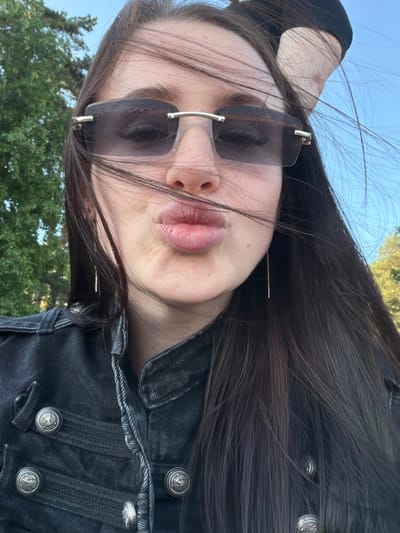 A close-up selfie features a young woman outdoors in Repino, Russia. Her dark brown, long hair is windswept, obscuring parts of her face. She wears rimless, dark rectangular sunglasses with silver-colored metallic accents at the temples, covering her eyes. Her lips are pursed in a pouting expression. Freckles are visible on her nose and cheeks. She wears a black denim jacket with a structured collar, featuring multiple decorative silver-colored buttons embossed with a heraldic-style emblem, possibly a double-headed eagle. Long, thin gold-colored threader earrings with a small bar at the end are visible on both sides. A black fabric item, possibly a headband or cap, is partially visible at the top right of her head. The background consists of dense green tree foliage on the left and a clear blue sky on the right, indicating an outdoor, breezy environment.