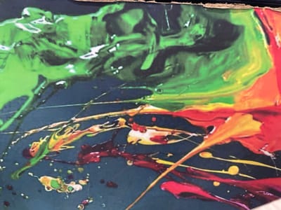 The image shows a close-up of an abstract painting with vibrant colors. Prominent colors include green, black, red, orange, and yellow. The paint appears to be applied in drips, splatters, and streaks, creating a chaotic and dynamic visual effect. A dark blue or black background is visible between the colored elements. There are no recognizable people or objects depicted in this image. There is no indication of any location in the city of Karshi, Nigeria.