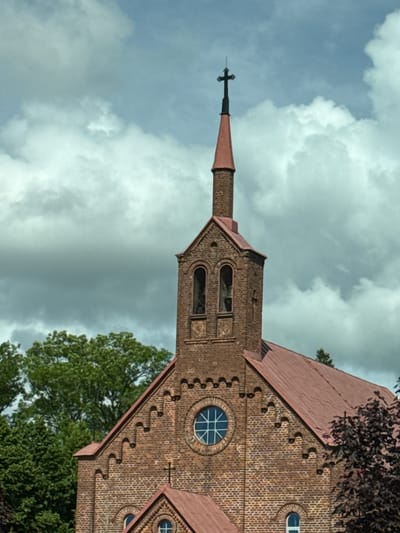 The image shows the exterior of a brick church with a red, sloped roof. A tall, slender spire with a cross on top rises from the church. The spire is made of brick and topped with a conical, copper-colored roof. Below the spire, the church has a bell tower with two arched openings. A circular window with a cross-shaped mullion is visible on the main body of the church. The church is surrounded by green trees and a cloudy sky. This is the Holy Trinity Catholic Church in Šilutė, Lithuania.