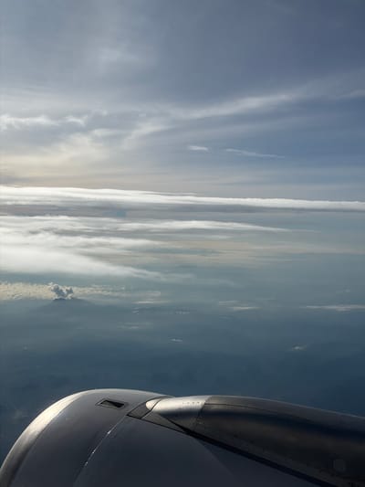 An aerial view from an airplane window captures a vast expanse of sky filled with layered clouds. Below the clouds, a hazy landscape unfolds, revealing mountains and a plume of smoke rising from a volcanic peak. In the foreground, the metallic engine of the aircraft is visible, with its curved housing and riveted details. The scene is characterized by soft, diffused light filtering through the clouds, creating a serene and expansive atmosphere.