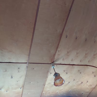 A single light fixture hangs from a wood-paneled ceiling. The ceiling is composed of light-colored plywood sheets with visible grain patterns, joined by thin wooden strips. Small, dark circular imperfections, likely knots or nail heads, are scattered across the plywood. The light fixture itself features a cylindrical white component at the top, from which a dark rod extends down to a translucent, amber-colored glass shade. The shade has an uneven, flared rim. The overall environment suggests an interior space with a rustic or unfinished aesthetic. The image is taken from a low angle, looking upwards at the ceiling. No people are present in the scene.
