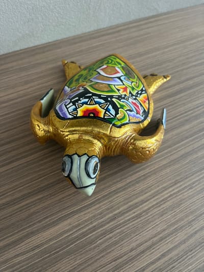 A single decorative turtle figurine is situated on a wooden surface, oriented with its head facing towards the lower left of the frame, in an indoor setting in Galatina, Italy. The figurine is primarily golden, featuring a textured, slightly wrinkled appearance across its body, flippers, and head. Its carapace is intricately decorated with a mosaic of colorful patterns, including vibrant greens, blacks, reds, yellows, purples, and whites, interspersed with gold accents and outlined details. These patterns combine geometric shapes with stylized organic and floral motifs. The turtle's head is a lighter, pale beige color with distinct dark grey and white painted eyes. The underlying wooden surface exhibits prominent parallel grain lines, and a plain, light-colored wall serves as the background. No people, active movements, or direct interactions are visible.