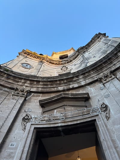 The image shows a section of a building in Acireale, Italy, shot from a low angle, looking upwards. The building is constructed from stone and features ornate architectural details, including curved moldings, carved embellishments, and a clock on the side of the building. The façade also includes a rectangular window or doorway, which is partially visible. A silver ladder is affixed to the side of the building near the window. The number "94" is written in white on the building's exterior. The sky is visible behind the building and is a clear, pale blue color.