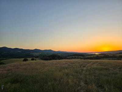 This is a landscape photograph taken at sunset in Golyama Zhelyazna, Bulgaria. The scene depicts rolling hills covered in tall grass in the foreground, with a treeline and more distant, darker hills forming the midground. A body of water, likely a lake or river, is visible in the far distance. The sky is a gradient of colors, transitioning from a pale blue at the top to a vibrant orange and yellow near the horizon, indicating the sun is setting. The light is soft and warm, casting long shadows. There are no people or man-made objects prominently featured, emphasizing the natural beauty of the rural setting. The overall mood is peaceful and serene, characteristic of a quiet evening in the countryside. There is no visible text in the image.