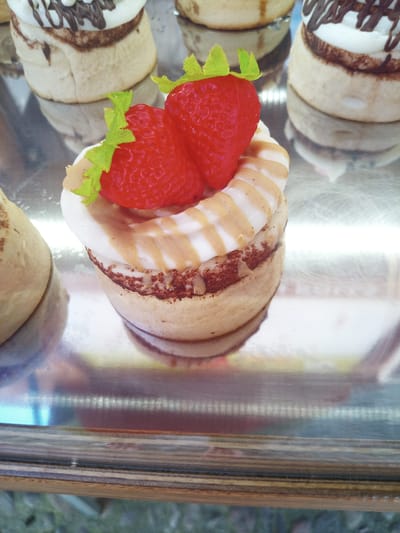 This is a close-up, eye-level, vertical shot of two artificial strawberries with green plastic leaves on top of a pastry. The pastry is a round cake with white frosting drizzled with a tan caramel-like sauce. The cake itself appears to be layered with a darker brown filling or frosting between two lighter cake layers. The pastry sits on a reflective silver tray. The background shows other pastries, also on a reflective surface. The scene is indoors, likely in a bakery or display case. The location is stated to be in Besalú, Spain. There are no people visible in the image.