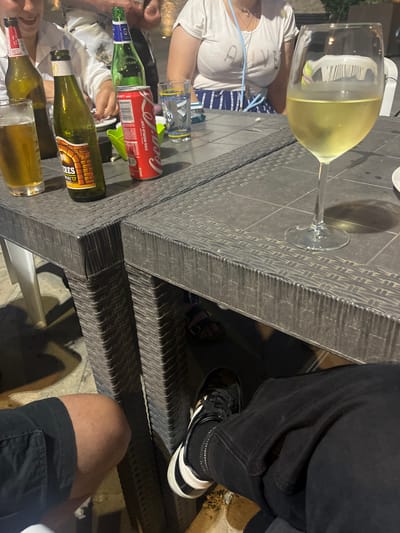 The image depicts a group of people seated around a dark, textured table, likely outdoors given the lighting and the presence of vegetation in the background. The time of day appears to be evening or night, suggested by the artificial lighting and the darkness of the surroundings.
The table is laden with drinks, including a glass of white wine, bottles of what appear to be beer and a can of Coca-Cola with "FIFA World Cup" visible text. The people are casually dressed, with one person wearing a white t-shirt and another in black pants and sneakers. One individual is wearing a blue and white striped skirt.
The scene conveys a relaxed atmosphere, suggesting a social gathering, possibly a dinner or drinks at a restaurant or bar. The focus is primarily on the table and the drinks, with glimpses of the people enjoying themselves. The overall impression is one of leisure and socializing. The location is identified as Vallo della Lucania, Italy.