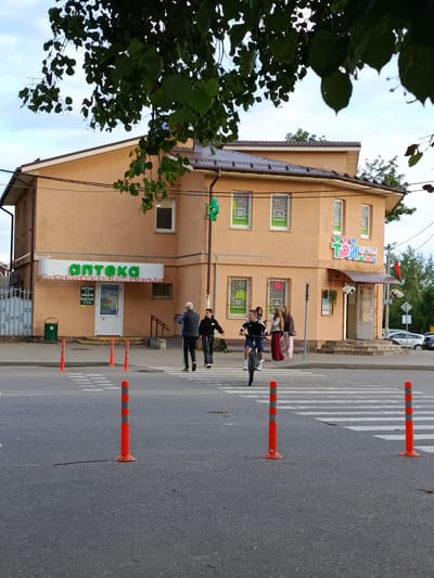 An exterior, eye-level, medium shot captures a street scene in front of a two-story commercial building. The building, painted a peach color, has a dark roof and multiple windows. A prominent green sign reading "аптека" (pharmacy) is visible on the left side of the building. On the right side, a colorful sign displays "ТРИ ЦЕНЫ" (three prices). Several people are present: a man in a blue jacket and black pants, a boy in a black hoodie, a girl in a white hoodie and pink pants, and another girl in a brown hoodie and maroon pants. A young man is riding a bicycle across a pedestrian crossing in the foreground. Orange and gray striped traffic cones are placed on the road, delineating the pedestrian crossing. The sky is cloudy. A lush green tree hangs over the top of the building, with its leaves obscuring a portion of the sky. The location is the city of Смілавіцкі сельскі Савет, Belarus.
