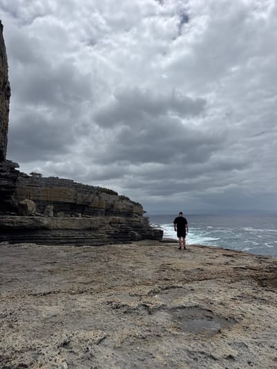 A figure stands on a rocky edge overlooking the ocean under a cloudy sky. The rock formation features layered strata, adding texture to the scene. A small puddle glistens on the flat, textured surface of the cliff where the figure stands. The sea stretches towards the horizon, its color mirroring the overcast sky above.