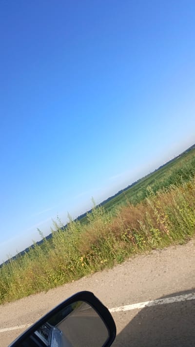 The image shows a roadside view of a rural landscape under a clear blue sky. A car's side-view mirror is visible in the lower left corner, reflecting a portion of the road and the car's body. The road appears to be unpaved or gravel. To the right of the road, there is a field of tall, dry grasses and some greenery, with a more densely vegetated area in the distance. The horizon line is low, with the vast expanse of the sky dominating the upper portion of the image. In the far distance, silhouetted against the sky, are what appear to be industrial structures or smokestacks. The tilt of the image suggests it was taken from a moving vehicle. The location is identified as Zuy, Russia, implying a rural or semi-rural setting within or near that area. There are no people or animals visible in the scene.
