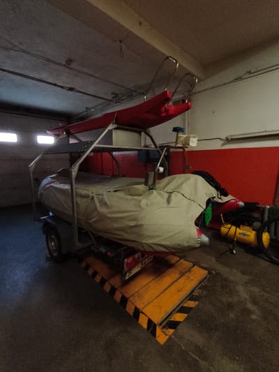 In a garage in Tukums, Latvia, a trailer is loaded with a boat covered by a tan tarp. Above the boat, two red kayaks are secured to a metal rack. A yellow air compressor with the brand name "FIAC" is visible to the right of the trailer. The garage has a concrete floor and red walls. Two windows with natural light are visible on the left side of the image. The scene appears to be a storage area for recreational equipment.