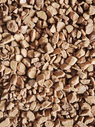 The image is a close-up, overhead view of a textured surface composed of numerous small, irregular, tan-colored granules. These granules appear to be coarse and somewhat porous, with varying shapes and sizes, suggestive of instant coffee granules or similar food product. The lighting creates subtle shadows, highlighting the three-dimensional nature of the material. There is no discernible background or environmental context visible. The image does not depict any location in the city of Kursk, Russia, nor does it contain any people, actions, or interactions.