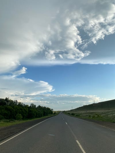 This is a forward-facing, eye-level shot of a paved road leading into the distance. The road has a white dividing line and a white line along the right shoulder. To the left of the road is a dense line of green trees. To the right, the land slopes upwards into a grassy hill with several power lines visible. The sky is a bright blue with large, dramatic white clouds, some appearing wispy and others more dense. The overall impression is a scenic drive in a rural or semi-rural area, possibly on the outskirts of a city given the presence of power lines and a road suitable for travel. The location is stated to be in Verkhneozyornoye, Russia, though there are no specific landmarks to confirm this within the image itself beyond the general landscape. There are no people visible, and the scene is static, depicting a moment of travel.
