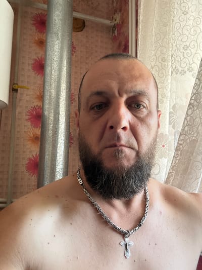 The image is a close-up, selfie-style portrait of a middle-aged man with a full beard and a goatee. He is bare-chested and wearing a thick, silver chain with a cross pendant. He has a serious expression and his eyes are looking directly at the camera. The background appears to be an indoor setting with patterned wallpaper depicting flowers, a silver pipe, and a lacy curtain. A yellow valve is visible on a pipe to the left of the man. The lighting suggests it is daytime, possibly near a window. The man's skin shows some texture and a few moles. The overall impression is that of a casual, personal photograph taken in a domestic environment. There is no visible indication of the specific location within the city of Zhirnov, Russia, in this image.