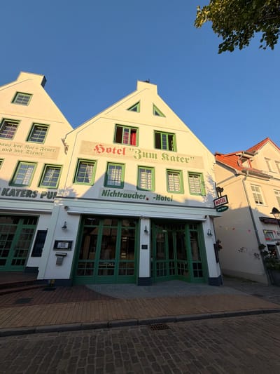 The image shows the exterior of a hotel in Rostock, Germany, identified by the signage "Hotel 'Zum Rater'" and "Nichtraucher - Hotel". The hotel appears to be in an older building with a distinctive gabled roof structure painted white, accented with green window frames and doors. The hotel sign, featuring a black cat silhouette, is mounted on the facade. The building is situated on a cobblestone street, with brick paving leading to the entrance. The sky is a clear blue. The time of day is late afternoon or early evening, as indicated by the long shadows cast by the building and a tree on the right side of the image. A few people are visible inside the hotel through the glass doors of the ground floor. The surrounding buildings also have traditional architectural styles.